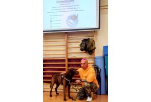 Prowadzący w żółtej bluzie klęczy przy swoim brązowym psie asystującym podczas warsztatów w sali gimnastycznej.