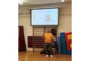 Prelegent prowadzi warsztaty  w sali gimnastycznej przy drabinkach. Za nim widać ekran z prezentacją, a obok niego leżącego psa asystującego.